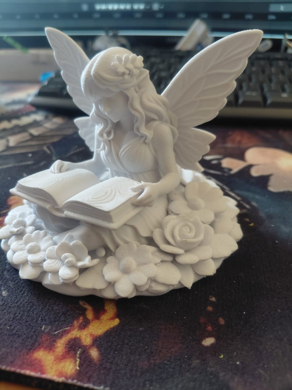 3D printed fairy statue reading a book, surrounded by flowers.