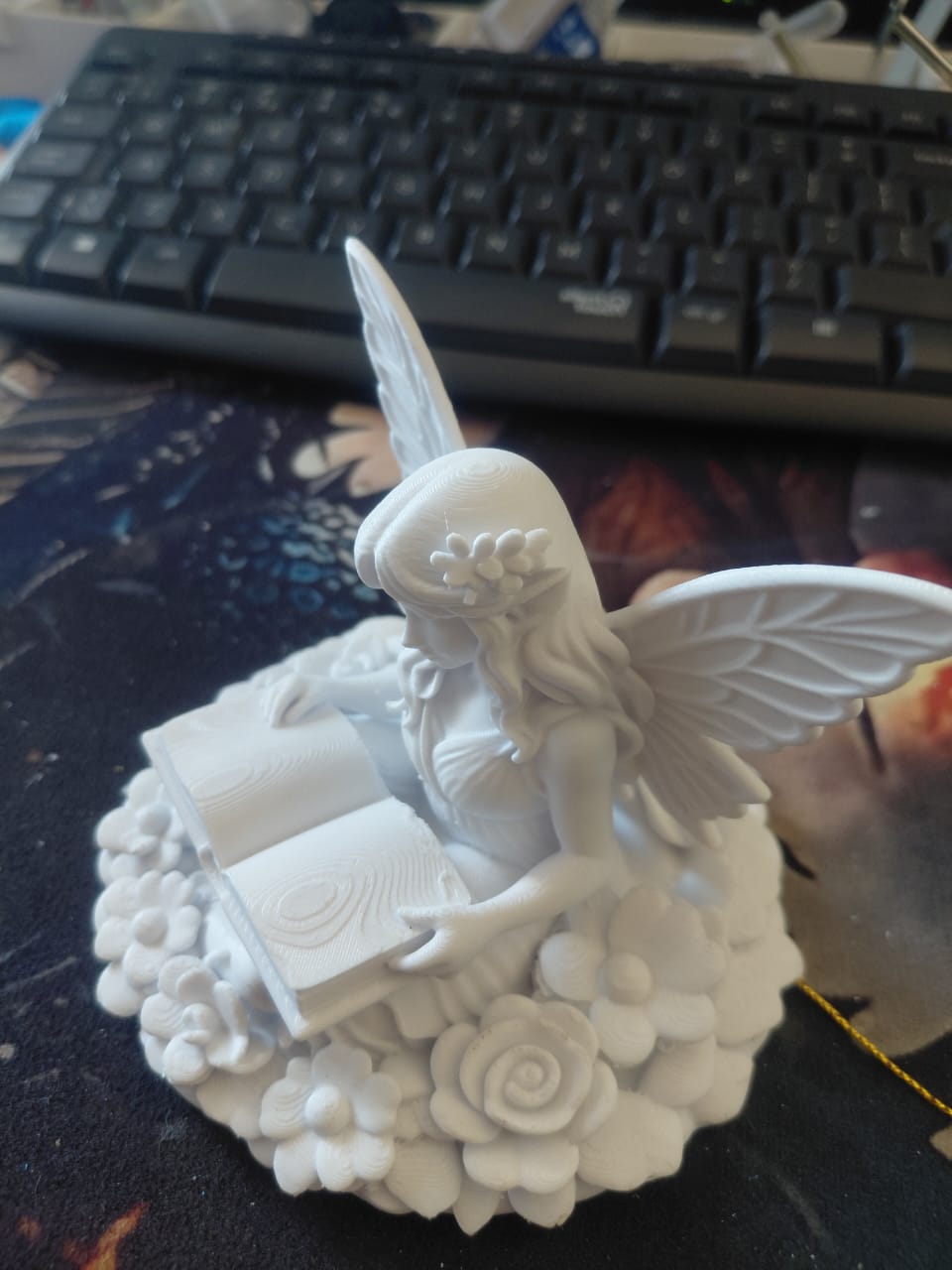 3D-printed fairy figurine reading a book, surrounded by flowers.