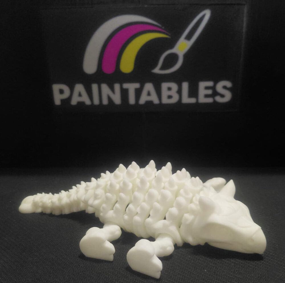 A white 3D printed Ankylosaurus skeleton lying flat in a resting pose. The view highlights the textured armored spikes along its back and the segmented articulated tail.