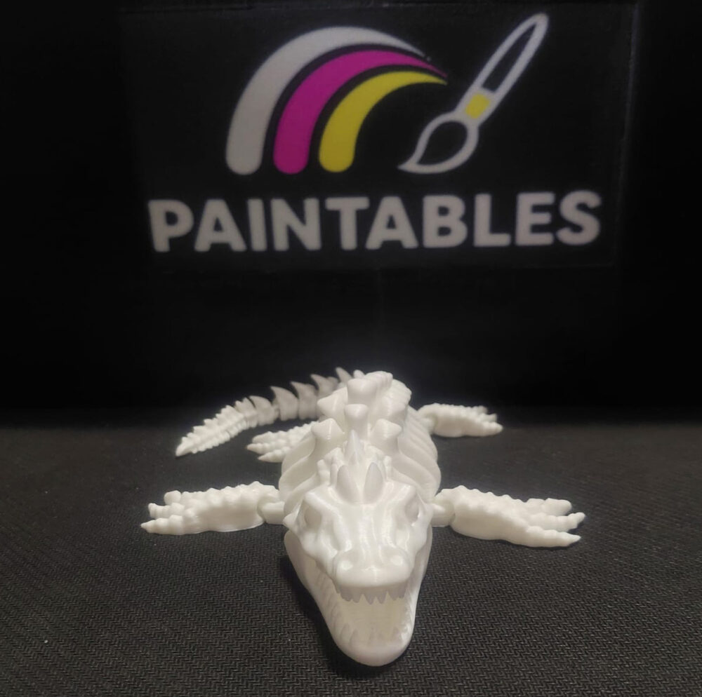 A head-on view of a white 3D printed Mosasaurus skeleton. The model features an elongated skull with a tapered snout, visible eye sockets, and four articulated flippers extended to the sides on a dark textured surface.