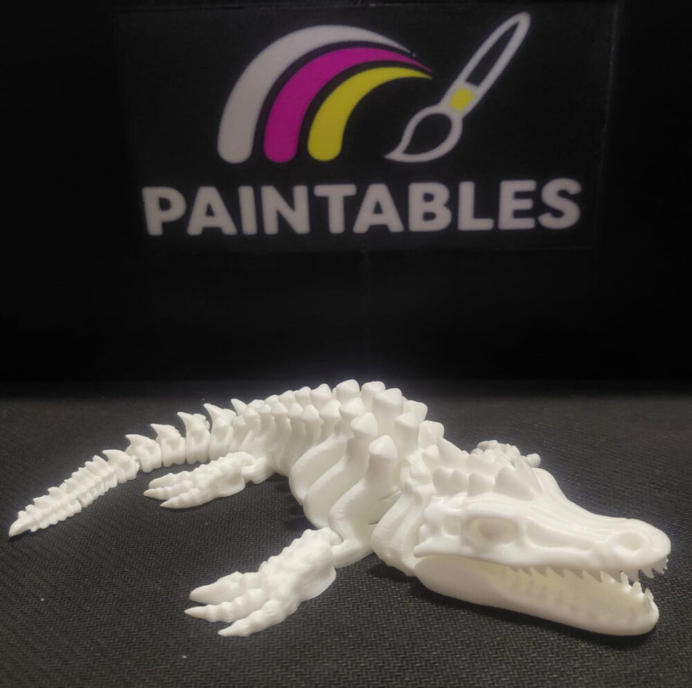 A white 3D printed Mosasaurus skeleton lying in a curved, swimming pose on a black surface. The model features a long, alligator-like skull with sharp teeth, four paddle-like flippers, and a segmented, flexible tail.