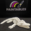 A white 3D printed Mosasaurus skeleton lying in a curved, swimming pose on a black surface. The model features a long, alligator-like skull with sharp teeth, four paddle-like flippers, and a segmented, flexible tail.
