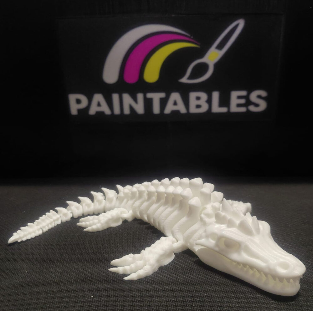 A white 3D printed Mosasaurus skeleton lying in a curved, swimming pose on a black surface. The model features a long, alligator-like skull with sharp teeth, four paddle-like flippers, and a segmented, flexible tail.