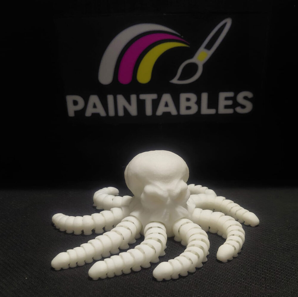 A white 3D printed articulated octopus named Ralph with a skull-shaped head. The model features eight thick, segmented, and highly flexible tentacles coiling outward on a black surface under a "Paintables" logo.