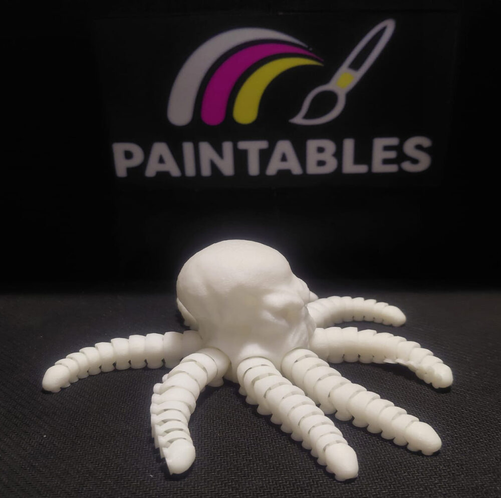 A side profile view of Ralph, a white 3D printed articulated octopus with a skull-shaped head. The model is shown on a black surface with its thick, segmented tentacles spread out, highlighting the intricate bone-like structure of the mantle.