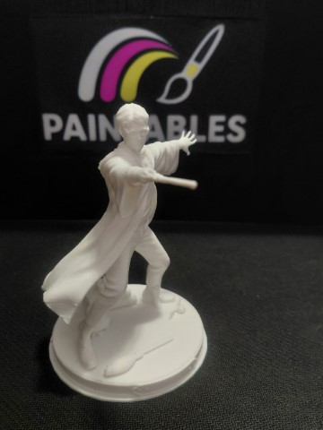 A white, unpainted 3D-printed miniature of Harry Potter in a dynamic spell-casting pose, standing on a circular base featuring a small broomstick and a golden snitch, set against a black "Paintables" background.