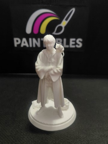 A white, unpainted 3D-printed miniature of Ron Weasley with a pet rat on his shoulder, standing on a circular base with a book and a cauldron, set against a black "Paintables" background.